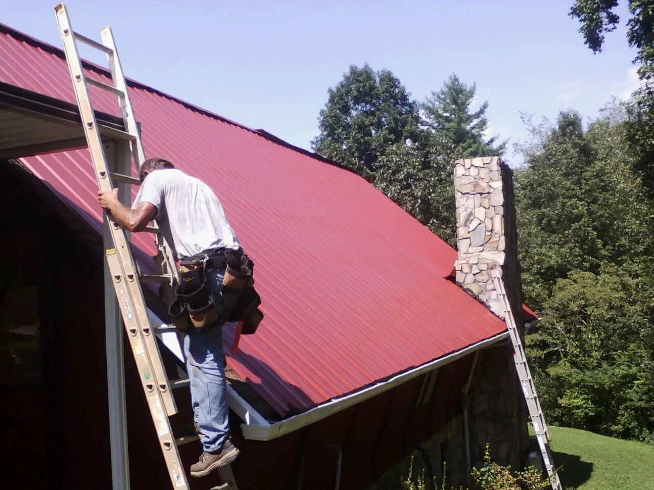 Roofer on a ladder beside a red metal roof and stone chimney — Soffit & Fascia Repair by Roofing Brothers in Winnetka